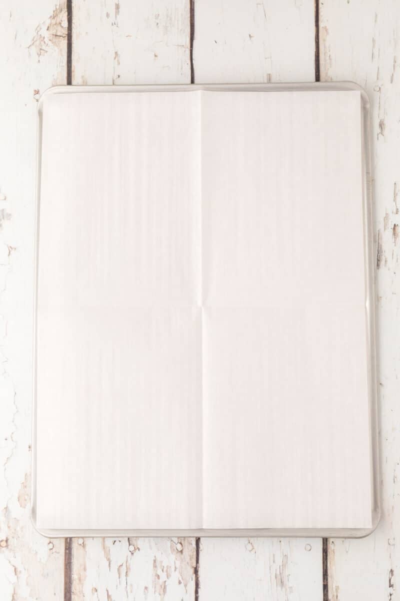 A square baking sheet lined with a large piece of white parchment paper on a rustic white wooden surface.