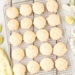 A cooling rack with 20 round, pale lime cookies sits on a white wooden surface, surrounded by halved limes, white flowers, and a yellow floral cloth.