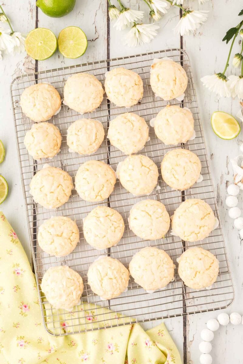 A cooling rack with 20 round, pale lime&nbsp;cookies sits on a white wooden surface, surrounded by halved limes, white flowers, and a yellow floral cloth.