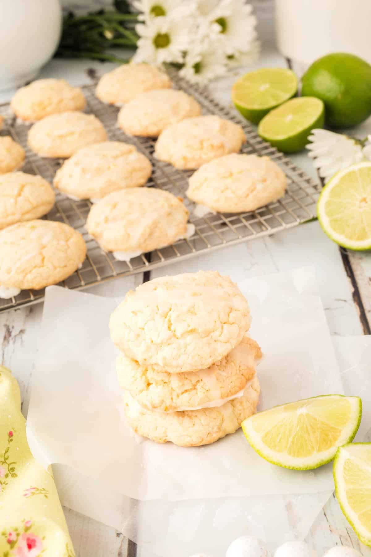 Three lime&nbsp;cookies are stacked in front of lime slices on parchment paper, with more cookies cooling on a rack in the background.