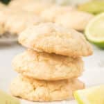 Three lime cookies stacked on parchment paper, with lime wedges and white candies nearby. More cookies and a blurred flower are in the background.