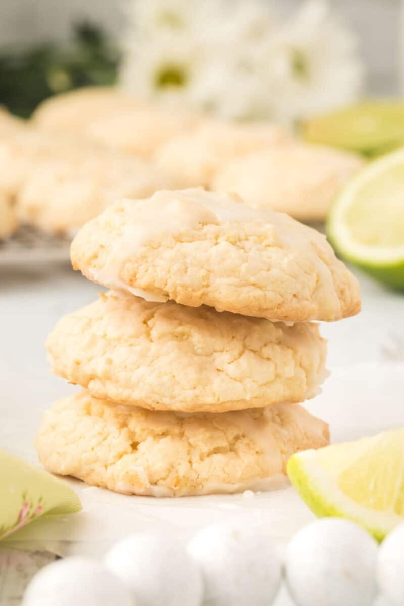 Three lime&nbsp;cookies stacked on parchment paper, with lime wedges and white candies nearby. More cookies and a blurred flower are in the background.