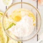 A glass mixing bowl containing flour, an egg, and wet ingredients sits on a white wooden surface next to a green cloth, eggshells, and a small bowl with zest.