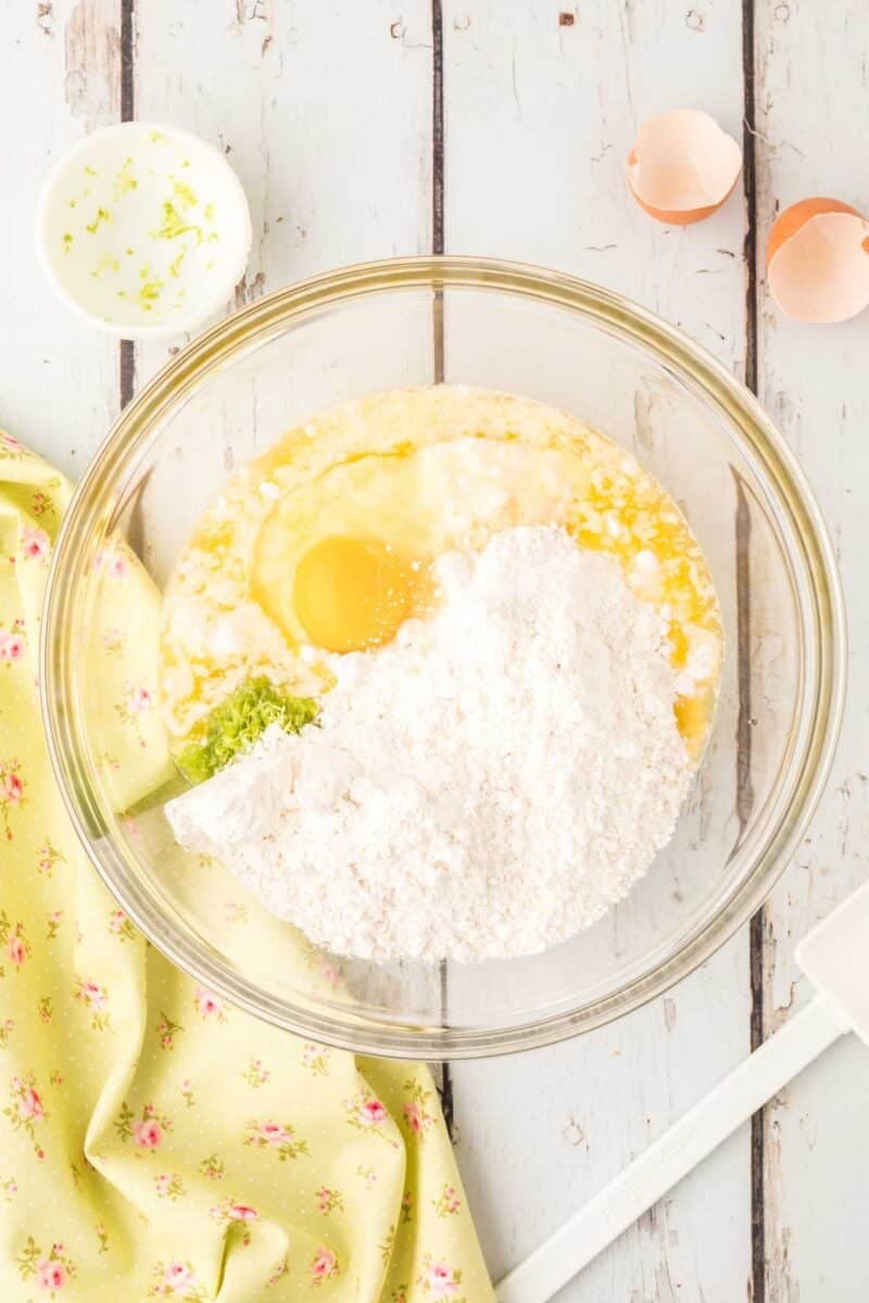 A glass mixing bowl containing flour, an egg, and wet ingredients sits on a white wooden surface next to a green cloth, eggshells, and a small bowl with zest.