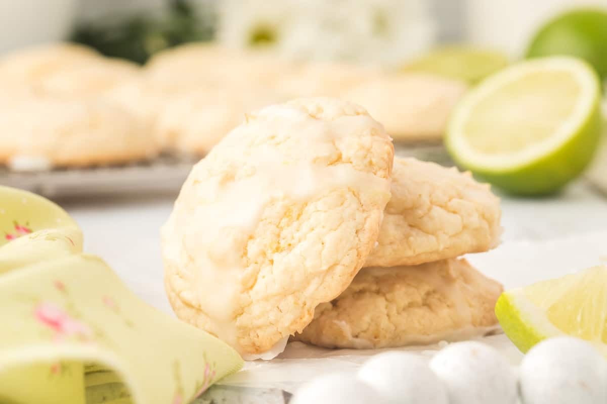 Two glazed lime&nbsp; cookies are stacked on parchment paper with a lime wedge and more cookies blurred in the background.