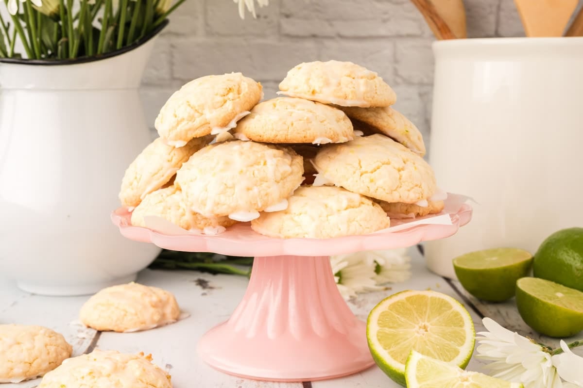 A stack of iced lime&nbsp; cookies sits on a pink cake stand, with sliced limes and white flowers in the background on a kitchen counter.