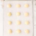 Twelve unbaked dough balls are arranged in rows on a parchment-lined baking sheet, with a cookie scoop and mixing bowl visible nearby.
