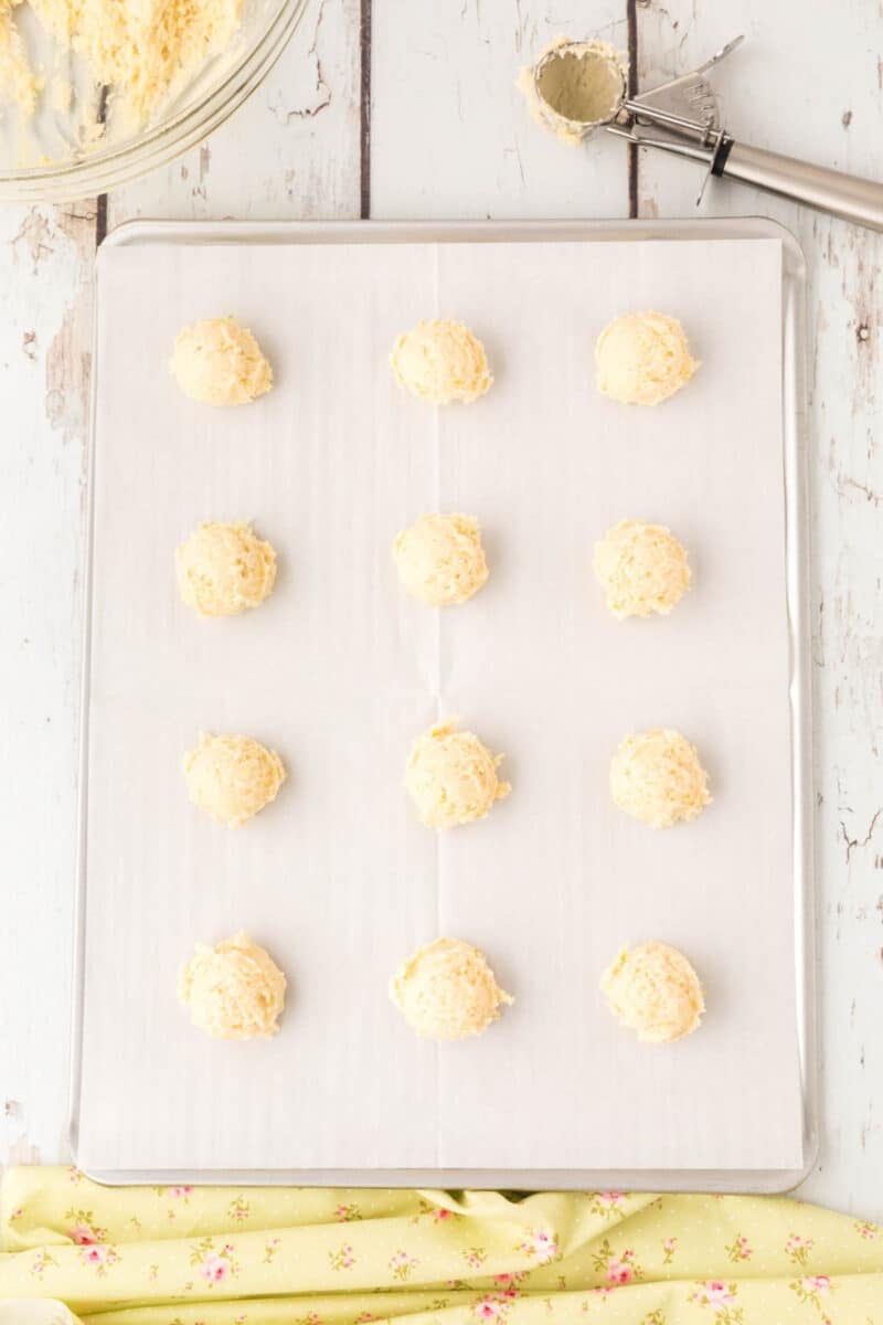 Twelve unbaked dough balls are arranged in rows on a parchment-lined baking sheet, with a cookie scoop and mixing bowl visible nearby.