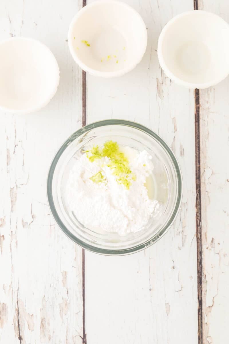 A glass bowl with powdered sugar and lime zest on a white wooden surface, with three empty small bowls above it.