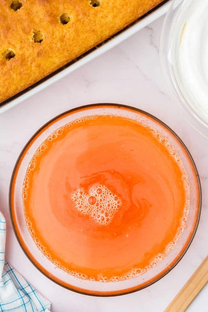 A bowl of orange gelatin mixture on a marble surface next to a partially visible poked cake and a bowl of whipped topping.