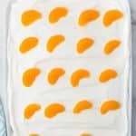 A rectangular dish filled with a layer of white whipped topping, decorated with evenly spaced mandarin orange segments on top. Orange slices and a napkin are visible in the background.