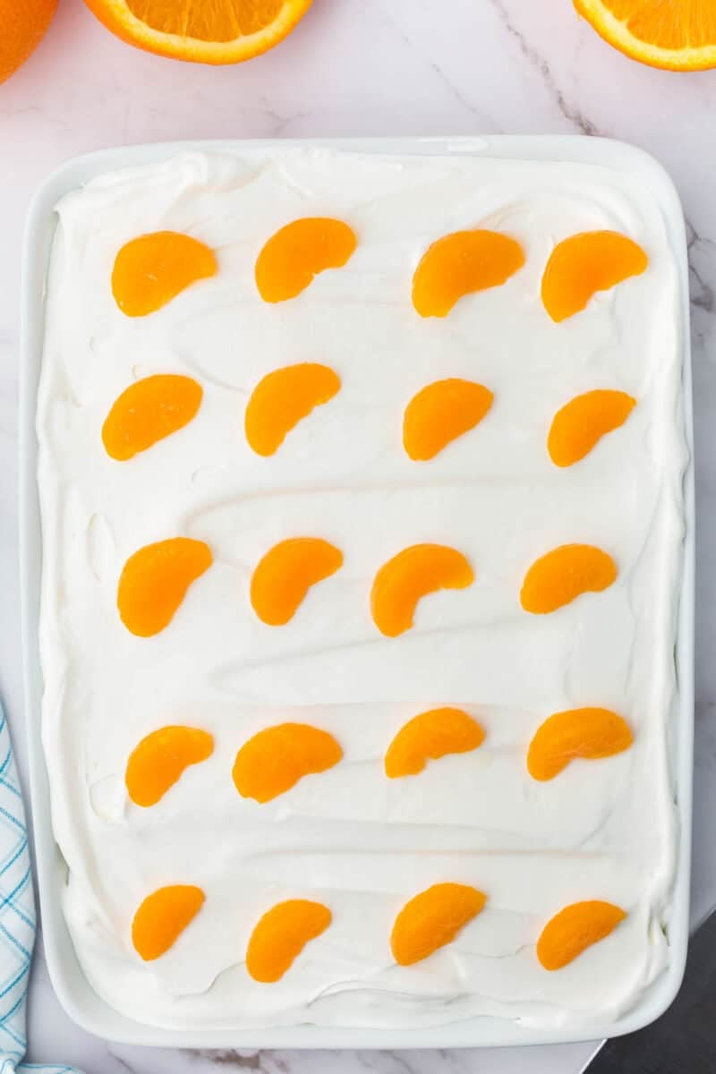 A rectangular dish filled with a layer of white whipped topping, decorated with evenly spaced mandarin orange segments on top. Orange slices and a napkin are visible in the background.