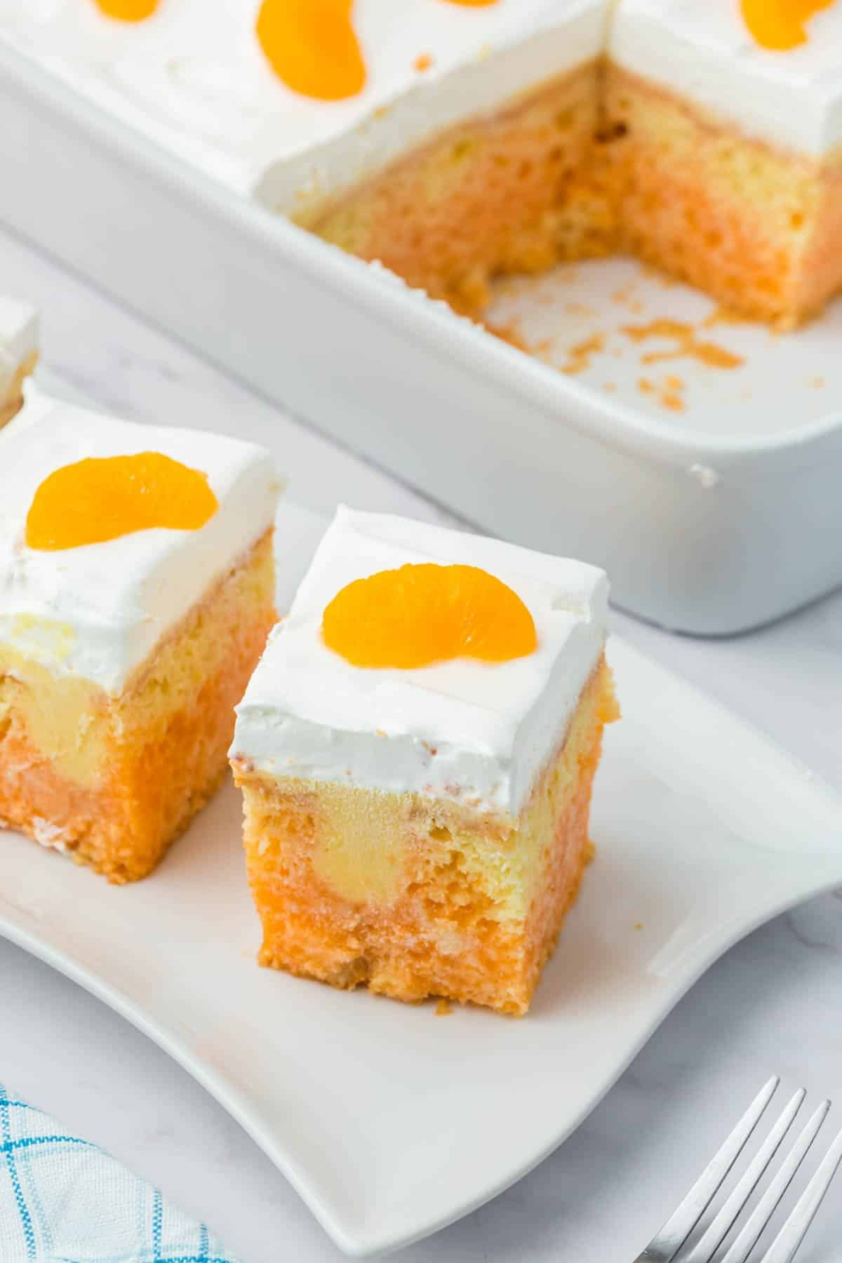 A white plate with two square slices of orange poke cake topped with whipped cream and a mandarin orange, with more cake in a white baking dish in the background.
