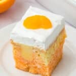 A square slice of orange and white cake topped with a layer of whipped cream and a mandarin orange segment, served on a white plate.