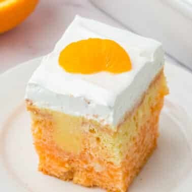 A square slice of orange and white cake topped with a layer of whipped cream and a mandarin orange segment, served on a white plate.