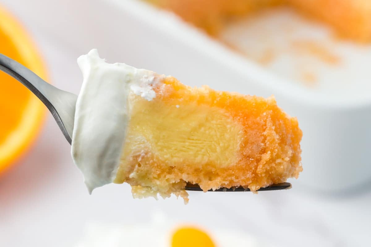 A close-up of a fork holding a slice of orange and yellow dessert topped with whipped cream, with a white dish in the background.