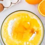 A glass bowl with eggs, flour, and wet ingredients being mixed, next to whole and halved oranges, egg shells, and a blue towel on a marble countertop.