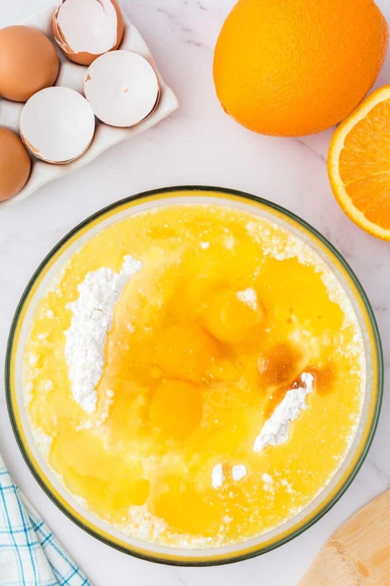 A glass bowl with eggs, flour, and wet ingredients being mixed, next to whole and halved oranges, egg shells, and a blue towel on a marble countertop.