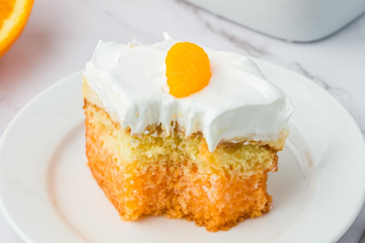 A slice of orange poke cake topped with white frosting and a mandarin orange segment, served on a white plate.