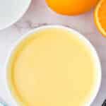 A white bowl filled with orange-colored batter sits on a marble surface next to a whole orange, a sliced orange, a white plate, and a whisk.