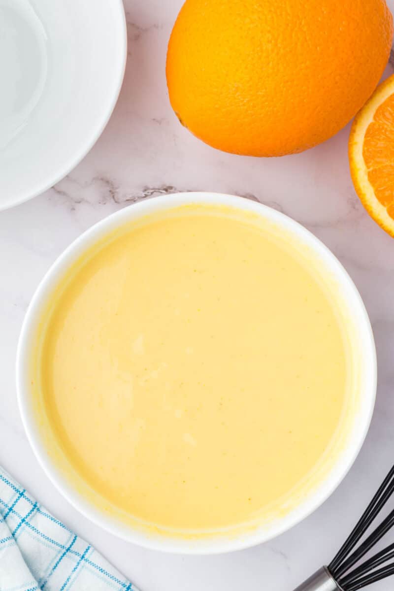 A white bowl filled with orange-colored batter sits on a marble surface next to a whole orange, a sliced orange, a white plate, and a whisk.