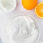 A clear bowl of whipped cream topped with powdered sugar sits next to a whole orange, a halved orange, and a small bowl of clear liquid on a white surface.