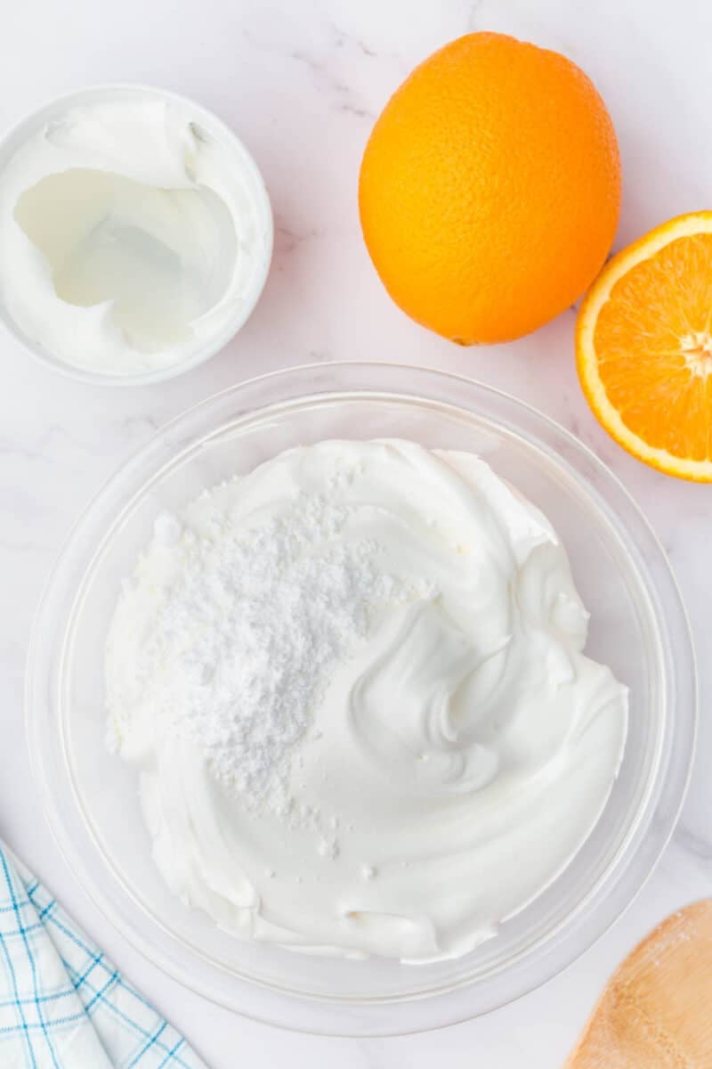A clear bowl of whipped cream topped with powdered sugar sits next to a whole orange, a halved orange, and a small bowl of clear liquid on a white surface.