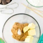 A glass bowl with peanut butter, butter, salt, and vanilla extract. Surrounding items include chocolate wafers, powdered sugar, a spatula, beaters, and wooden sticks on a white surface.