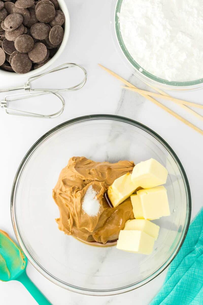 A glass bowl with peanut butter, butter, salt, and vanilla extract. Surrounding items include chocolate wafers, powdered sugar, a spatula, beaters, and wooden sticks on a white surface.