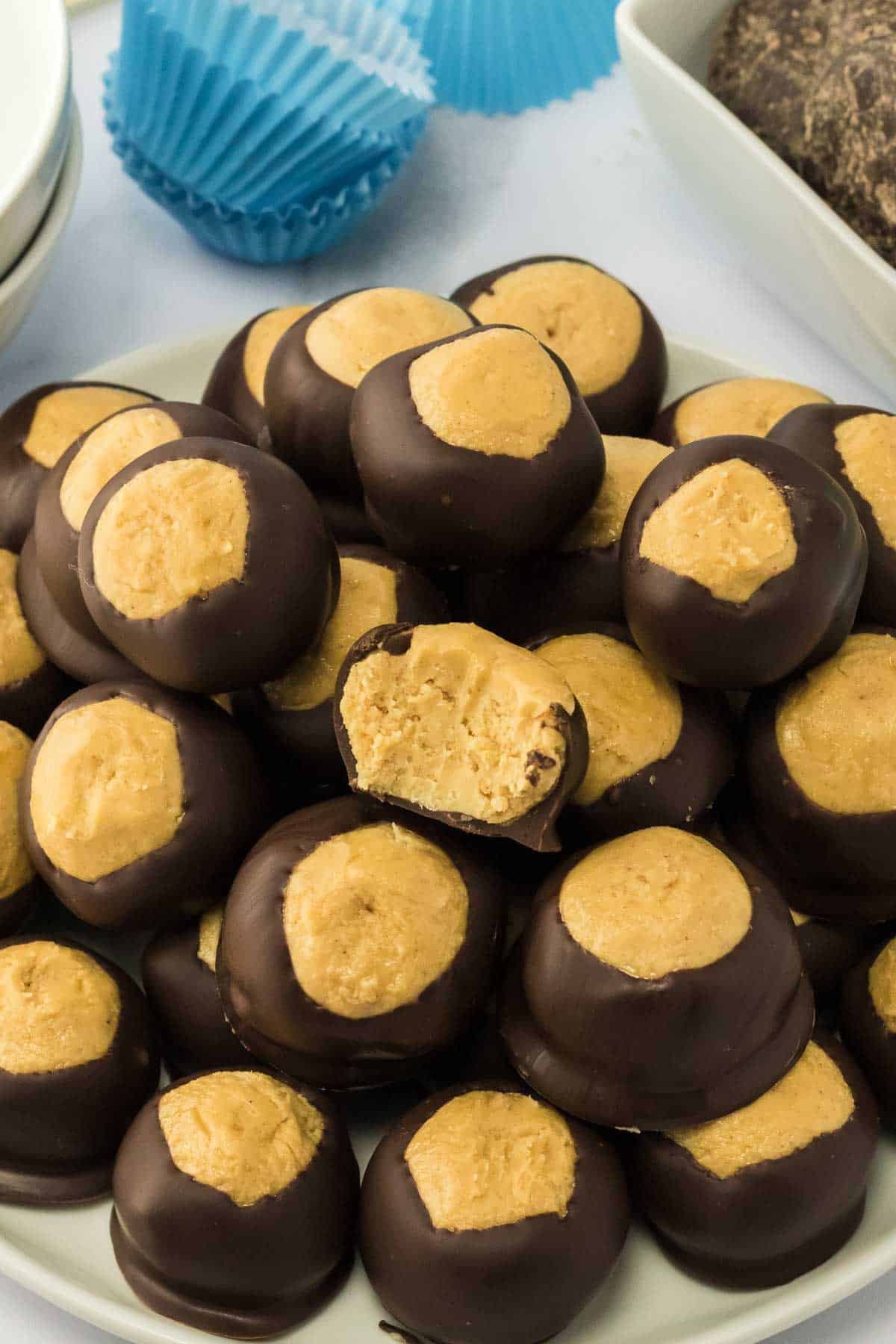 A plate of buckeye candies, featuring round peanut butter centers partially dipped in chocolate, with one cut open to show the inside. Blue cupcake liners are in the background.