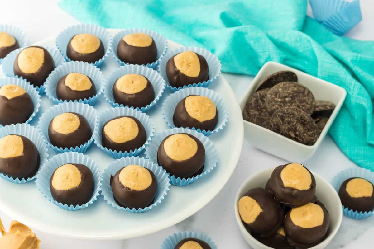 A plate and bowls of buckeye candies, with chocolate-coated peanut butter balls in blue paper cups, next to a bowl of chocolate wafers and a turquoise cloth.