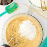 A mixing bowl with peanut butter mixture and powdered sugar being blended with a hand mixer, surrounded by chocolate wafers, powdered sugar, a spatula, and measuring tools.