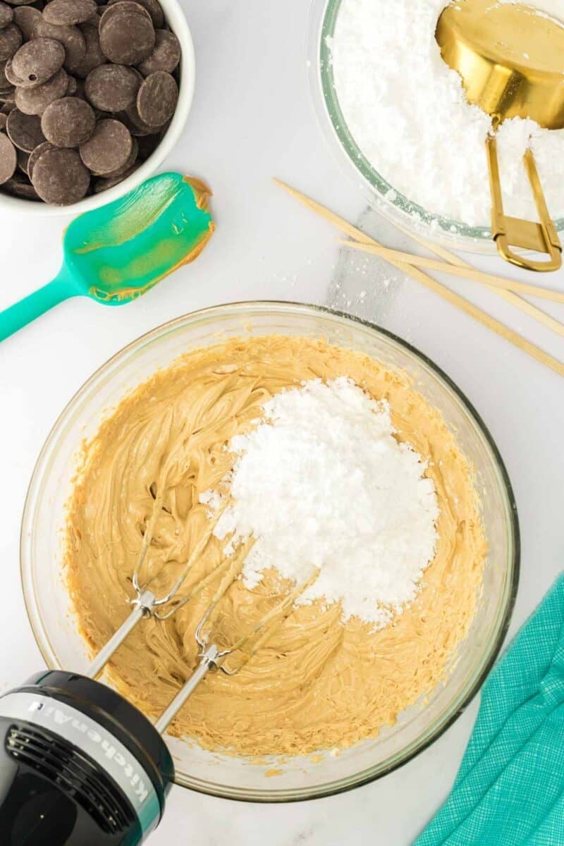 A mixing bowl with peanut butter mixture and powdered sugar being blended with a hand mixer, surrounded by chocolate wafers, powdered sugar, a spatula, and measuring tools.