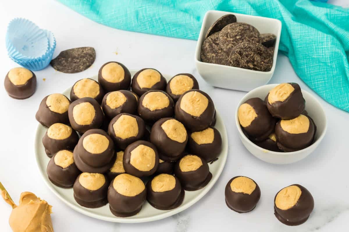 A plate and bowls filled with peanut butter balls partially dipped in chocolate, with extra chocolate pieces and a blue cloth nearby.