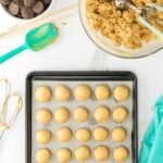 A baking tray with evenly spaced raw cookie dough balls, a bowl of cookie dough with a scoop, chocolate wafers, and baking utensils on a white surface.
