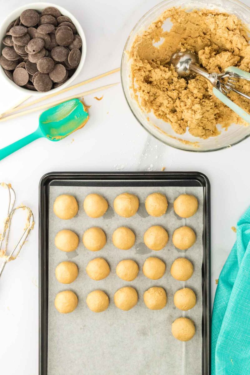A baking tray with evenly spaced raw cookie dough balls, a bowl of cookie dough with a scoop, chocolate wafers, and baking utensils on a white surface.