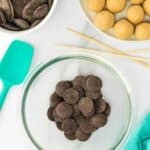 A glass bowl with chocolate melting wafers, a plate of dough balls, a bowl of more wafers, a teal spatula, wooden skewers, and a teal cloth on a marble surface.