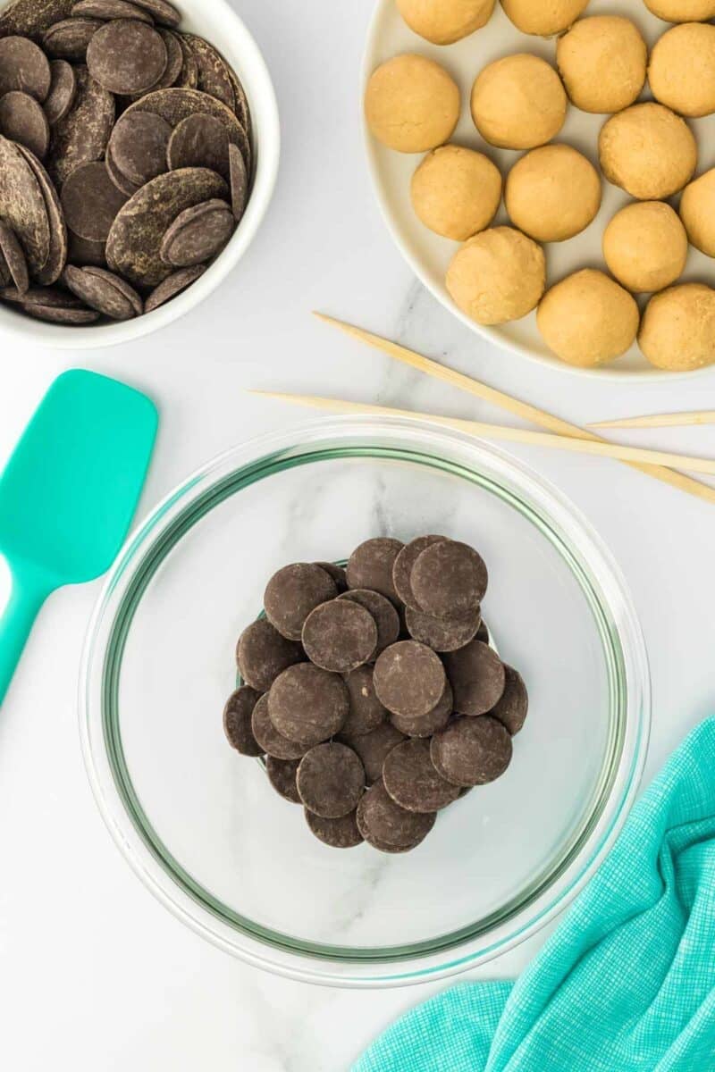 A glass bowl with chocolate melting wafers, a plate of dough balls, a bowl of more wafers, a teal spatula, wooden skewers, and a teal cloth on a marble surface.