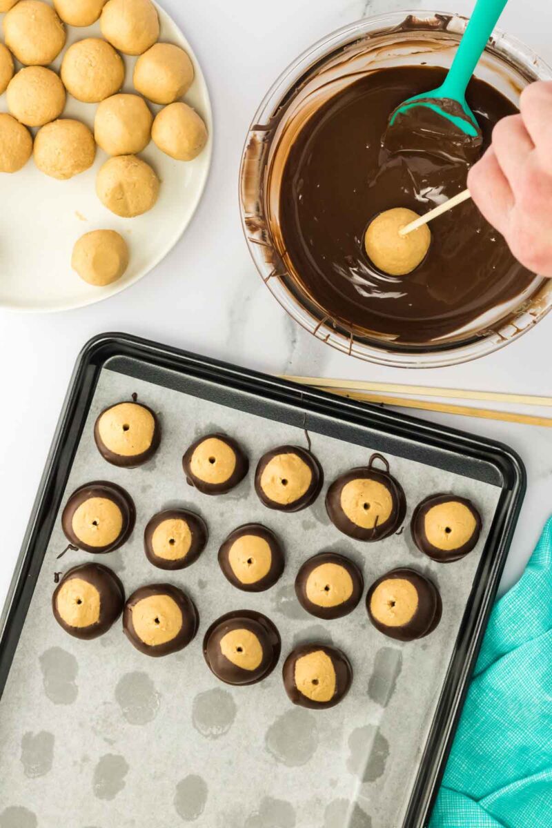 A hand dips a peanut butter ball into melted chocolate. Other dipped and undipped balls are on a plate and a parchment-lined baking sheet.