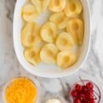 A white dish with halved, cored pears, next to small bowls containing shredded cheddar cheese, mayonnaise, and maraschino cherries on a marble surface.