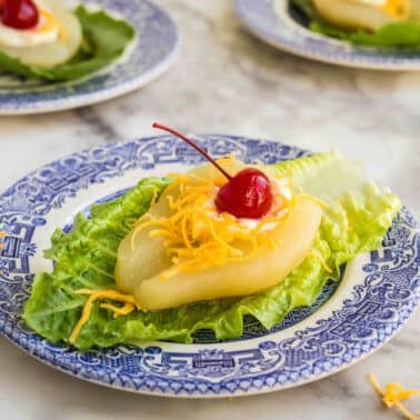 A halved pear on a leaf of lettuce, topped with shredded cheddar cheese, a dollop of mayonnaise, and a maraschino cherry, served on a blue and white plate.