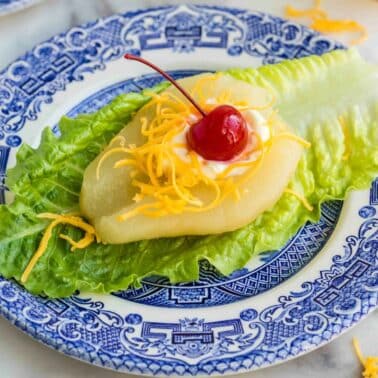 A lettuce leaf topped with a pear half, shredded cheddar cheese, mayonnaise, and a maraschino cherry on a blue and white patterned plate.