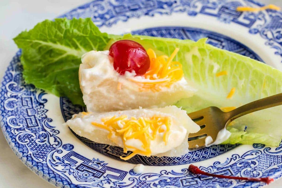 A wedge of iceberg lettuce on a blue and white plate, topped with mayonnaise, shredded cheese, and a maraschino cherry, with a fork holding a sliced piece.