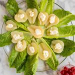 A glass plate with romaine lettuce leaves topped with canned pear halves, each filled with a dollop of mayonnaise. Bowls of shredded cheese and maraschino cherries are nearby.
