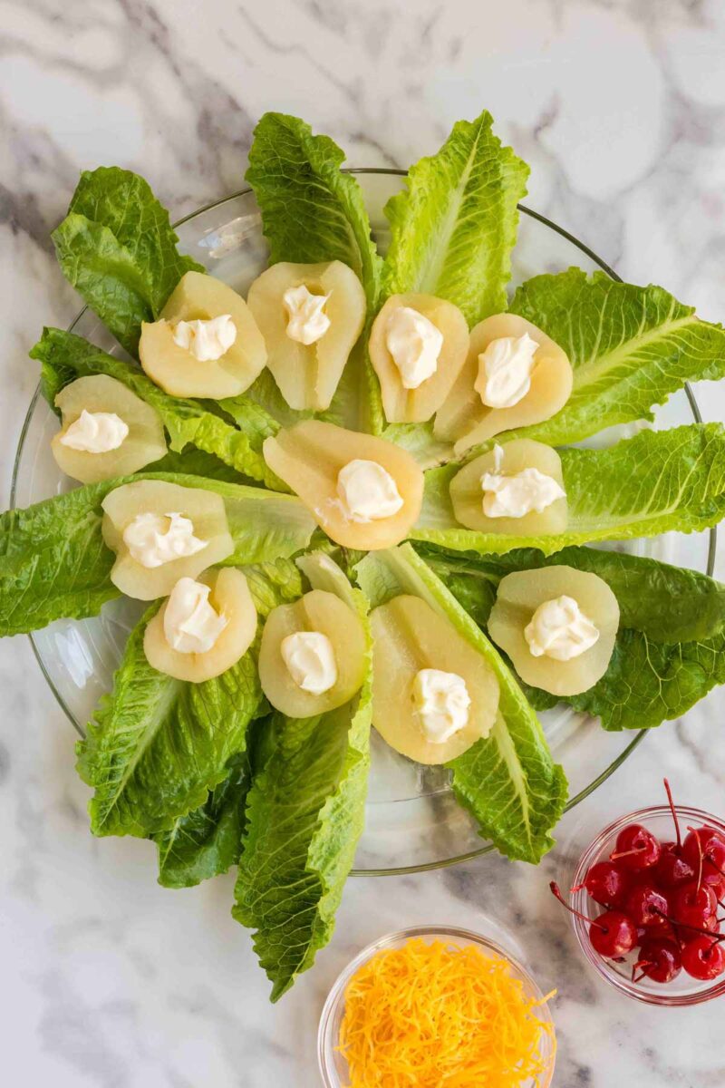 A glass plate with romaine lettuce leaves topped with canned pear halves, each filled with a dollop of mayonnaise. Bowls of shredded cheese and maraschino cherries are nearby.