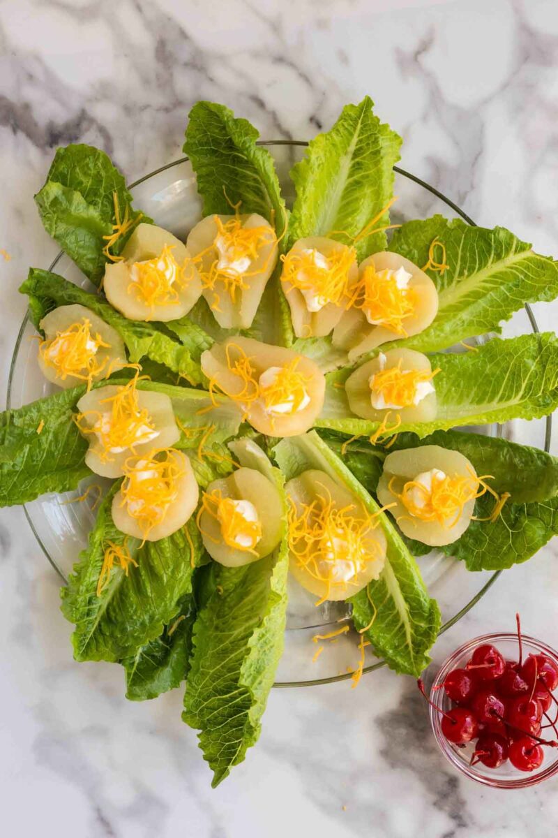 A plate of romaine lettuce topped with sliced pears, dollops of mayonnaise, and shredded cheddar cheese, with a bowl of maraschino cherries on the side.