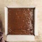 A square baking pan lined with parchment paper, filled with unbaked chocolate batter, sits on a light textured surface next to a purple cloth.