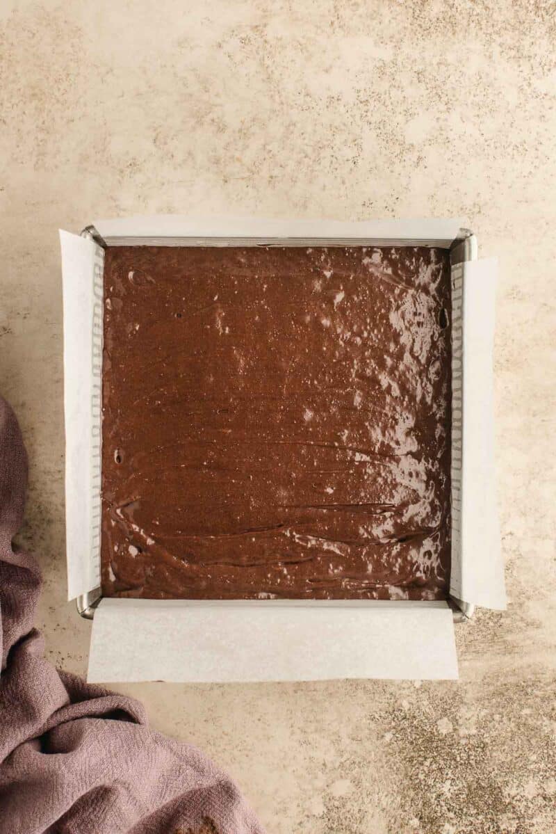 A square baking pan lined with parchment paper, filled with unbaked chocolate batter, sits on a light textured surface next to a purple cloth.