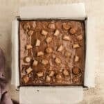 A square pan lined with parchment paper holds brownies topped with halved and whole miniature peanut butter cups.