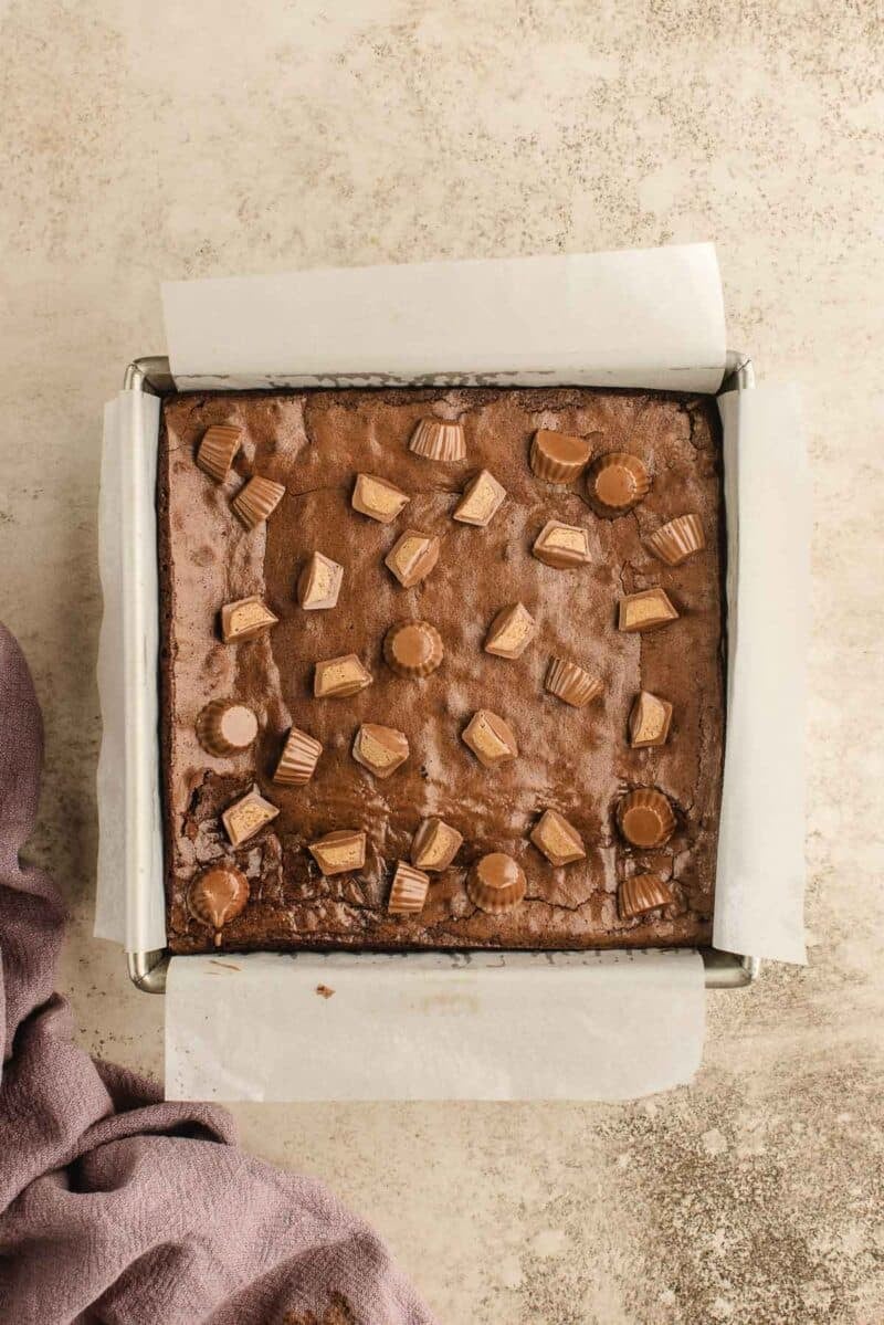 A square pan lined with parchment paper holds brownies topped with halved and whole miniature peanut butter cups.